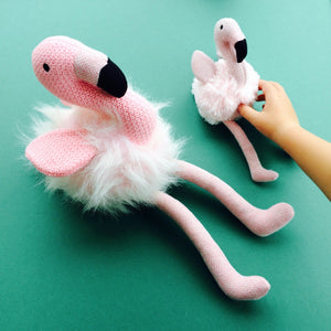 Large pink flamingo soft toy sitting next to flamingo rattle on green background with child's hand.